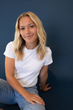 Sioux City, Iowa photographer. Young blonde woman in white t-shirt and jeans sitting against a dark blue wall smiling.