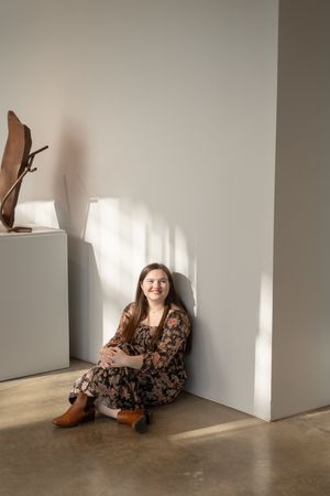 Sioux City, Iowa photographer. Smiling woman in floral dress sits on floor leaning against white gallery wall in sunlight.