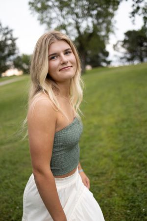 Sioux City, Iowa photographer. Blonde teen girl in green crop top and white skirt smiles softly in green park setting.