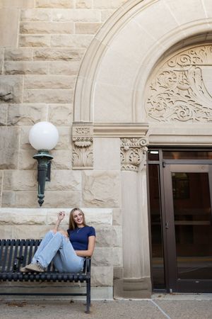 Sioux City, Iowa photographer. Smiling blonde woman in navy polo and jeans relaxing on bench outside ornate stone building entrance.