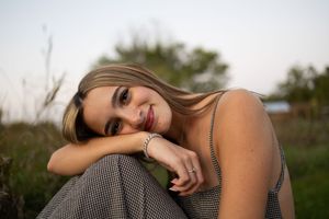Sioux City, Iowa photographer. Smiling blonde woman in checkered overalls resting chin on knees while sitting in green outdoor setting.