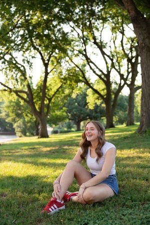 Sioux City, Iowa photographer. Laughing teenage girl sits on grass in park wearing white tee, denim shorts, and red Adidas.
