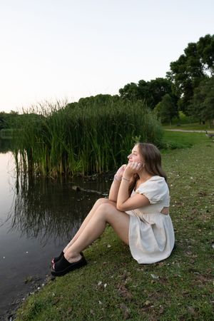 Sioux City, Iowa photographer. Young woman in white dress sits by peaceful lake shore surrounded by tall green reeds.