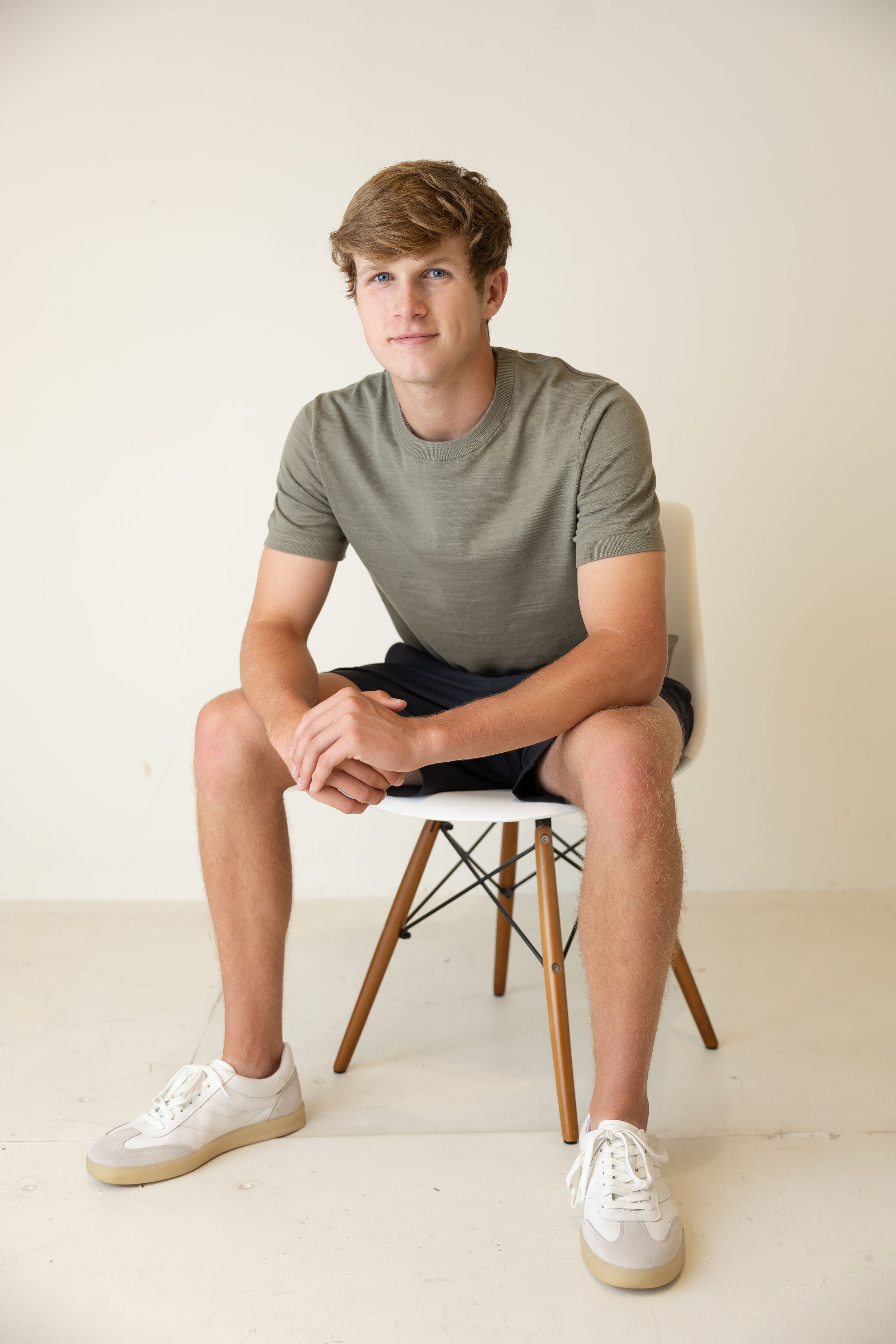Sioux City, Iowa photographer. Young man sitting on modern white chair with wood legs against neutral background.