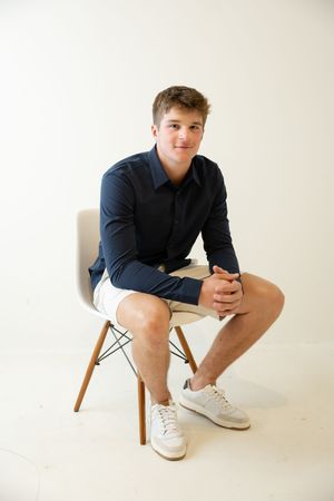 Sioux City, Iowa photographer. Young man in navy shirt and white shorts sitting on a modern chair against white background.