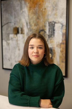 Sioux City, Iowa photographer. Young woman with brown hair in a green turtleneck sweater sitting with arms crossed before abstract wall art.