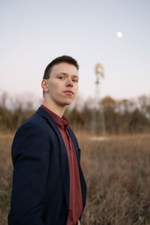 Sioux City, Iowa photographer. A young man in a navy blazer stands in a field at dusk with a windmill and moon behind him.