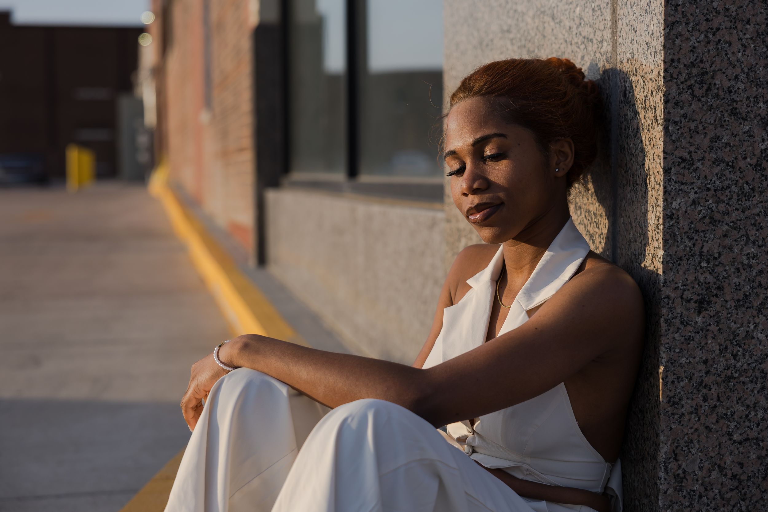 Sioux City, Iowa photographer. Young Black woman in white dress sitting against wall in golden sunlight.