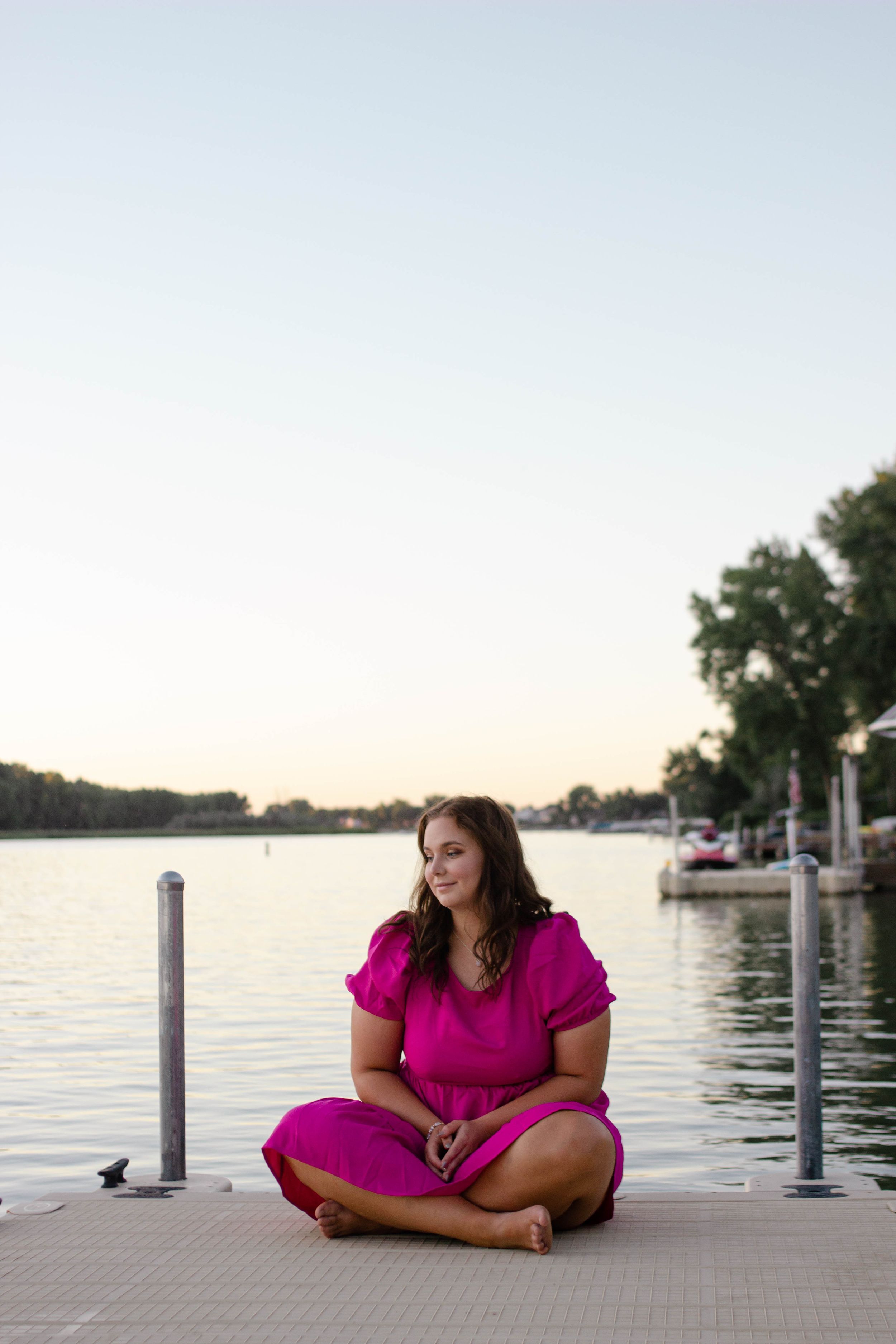 Sioux City, Iowa photographer. A young woman in a pink dress sits cross-legged on a dock by a calm lake at sunset.