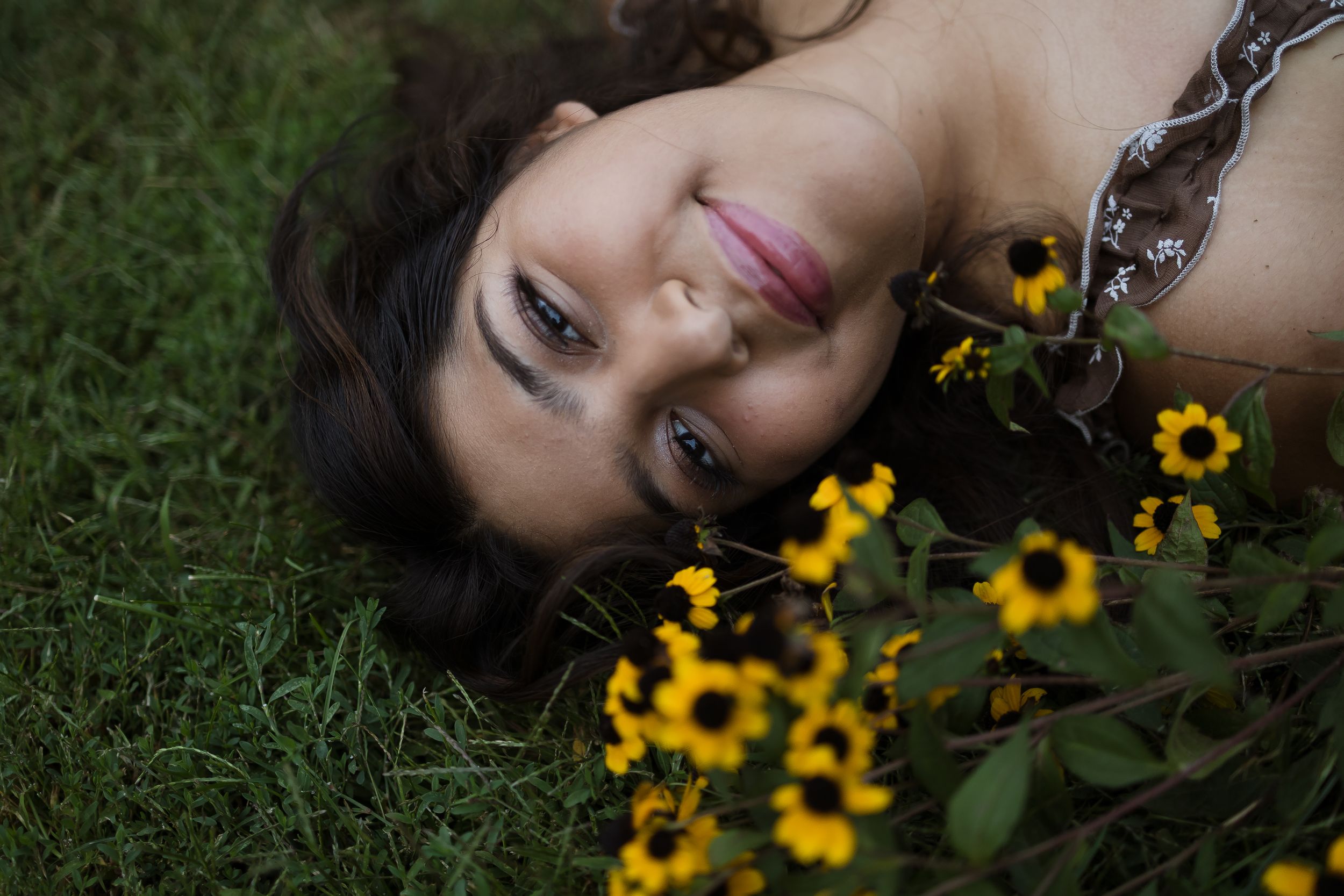 Woman lying on green grass smiling near yellow wildflowers in a serene outdoor setting.