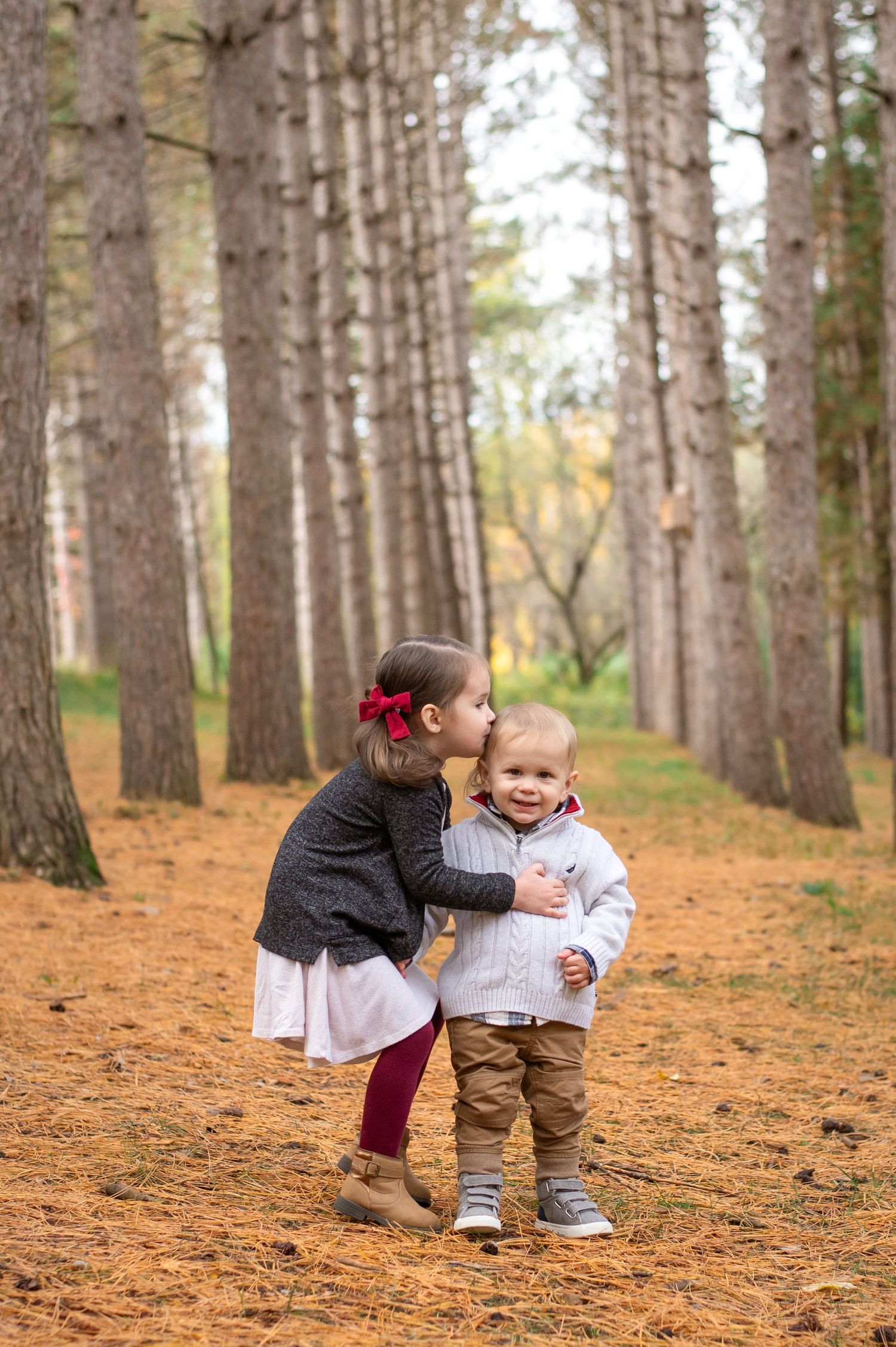 A couple sits together on a stone wall in matching green sweaters and jeans, sharing joyful laughter in a winter forest setting.