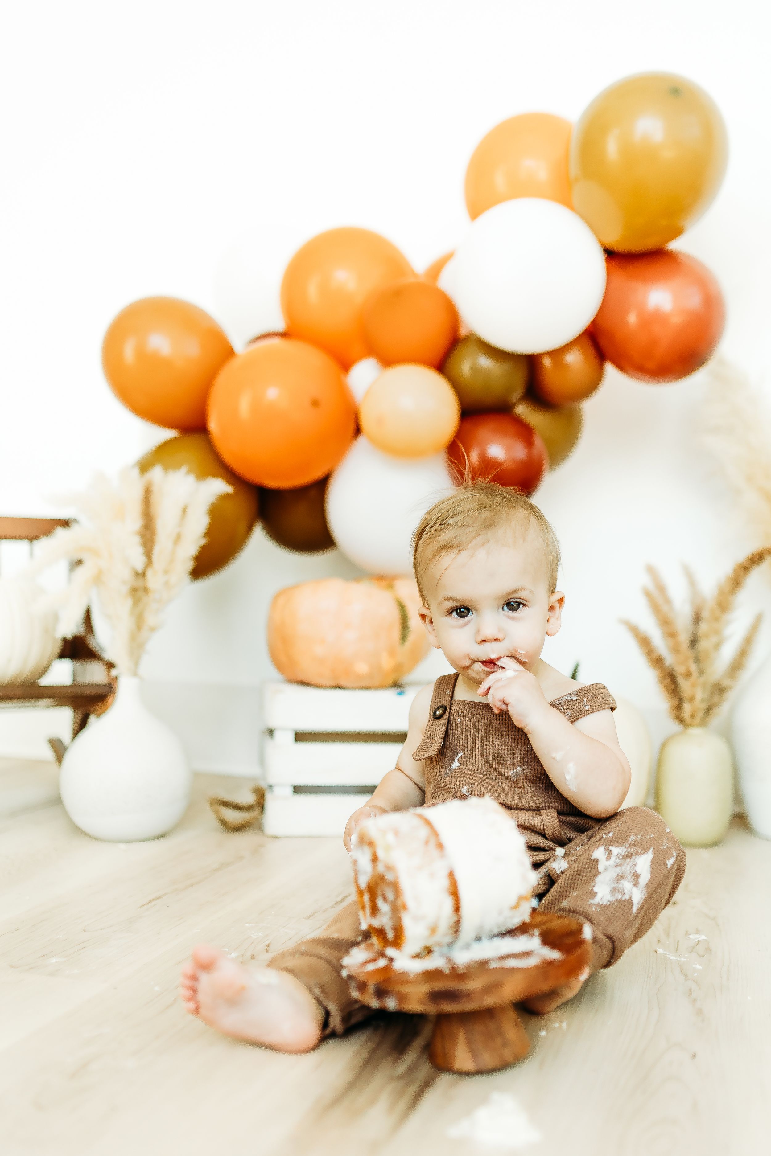 Festive autumn-themed party setup with orange and white balloon arch, wooden crate, and pampas grass decorations.