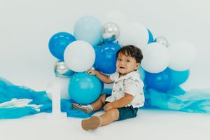 Baby wearing white outfit lies on floor surrounded by blue and white balloons in birthday cake smash photo session.