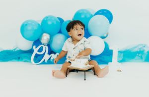 A baby sits on a white floor with blue balloon decorations and a small cake for a first birthday celebration.
