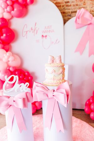 White gift boxes with pink bows arranged in a row with pink balloon decorations against a white and brick wall.
