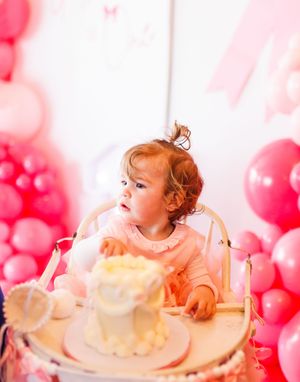 Baby with red hair enjoys a first birthday cake smash photo session with pink balloon decorations in the background.