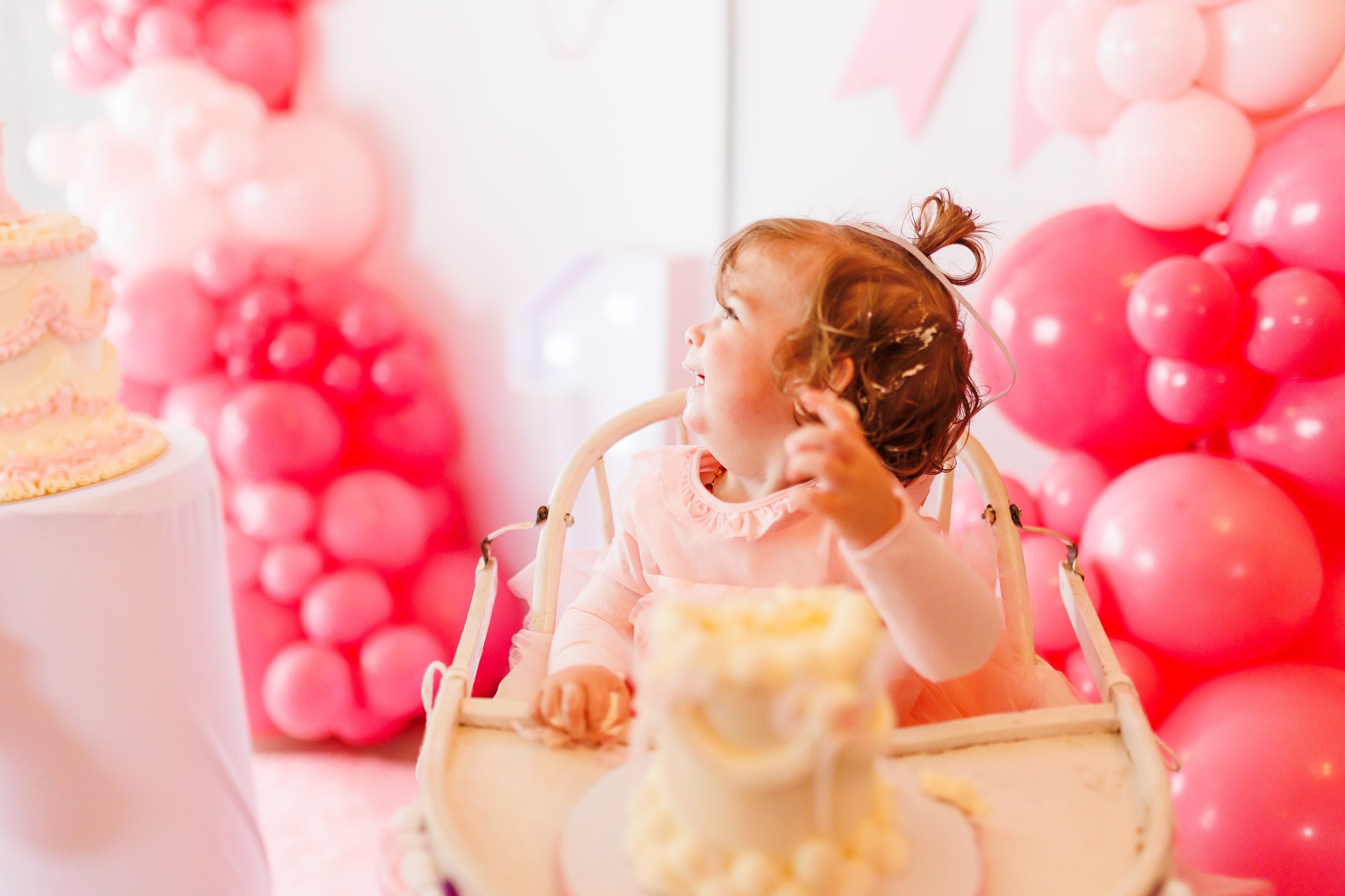 A baby in pink surrounded by pink balloon decorations leans over to blow out a birthday cake candle.
