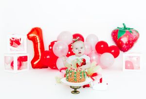 A festive holiday scene with pink and red balloons, gift boxes, and a decorative strawberry against a white background.