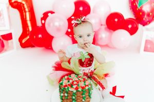 A festive first birthday cake smash setup with red and pink balloons, tulle dress, and decorated green cake.