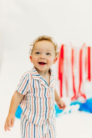 A baby wearing a striped red white and blue romper smiles joyfully against a bright festive background.