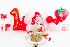 A cake smash photo setup with red balloon decorations, pink balloons, and strawberry themed party elements.