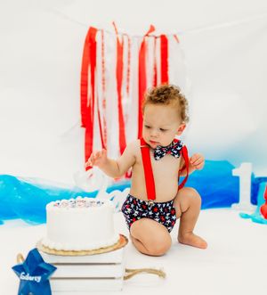 A first birthday cake smash photo setup with red streamers, blue accents, and a white cake against a light backdrop.