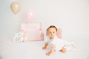 Baby girl in white dress sits near pink suitcase with balloons and \"One\" sign for first birthday photo.