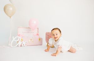 Baby girl in white dress crawling near pink gift box and balloons during first birthday cake smash photo session.