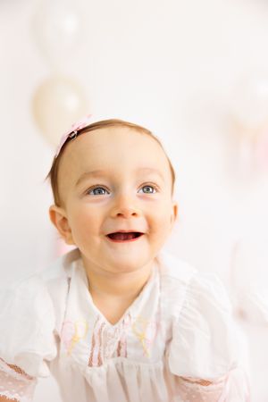Smiling baby girl in white dress with pink headband, looking up joyfully with balloons in soft background.