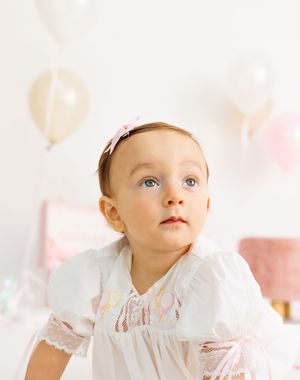 Baby girl in white lace dress with pink bow headband sits before pastel balloons at her first birthday party.