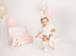 Baby in white outfit sits on bench with pink balloons and decorations celebrating a first birthday.