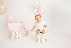 Baby girl in white outfit sits on bench with pink balloons and gift box at first birthday cake smash photo session.