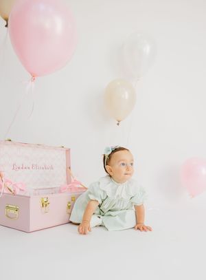 Baby girl in mint dress sits beside a pink personalized trunk with pastel balloons on white background.