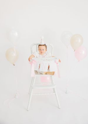 Baby girl in white dress sits in white high chair surrounded by pink and cream balloons for her first birthday celebration.