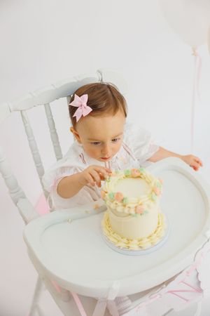 Baby girl in white dress with pink bow touches her first birthday cake in a high chair.