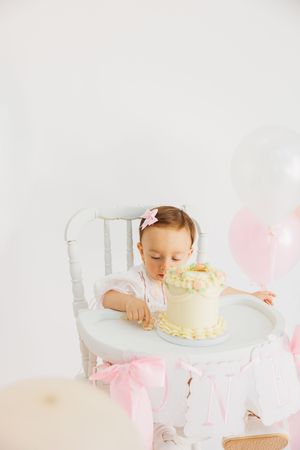 Baby girl in white dress sits in high chair with birthday cake and pink balloons on her first birthday.