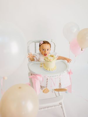 Baby girl in high chair celebrates first birthday with smash cake, pink bows, and balloons in white studio.