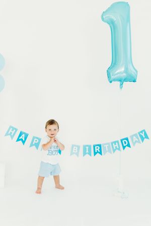 Baby in light blue outfit sits on white backdrop with turquoise number one balloon and birthday banner decorations.
