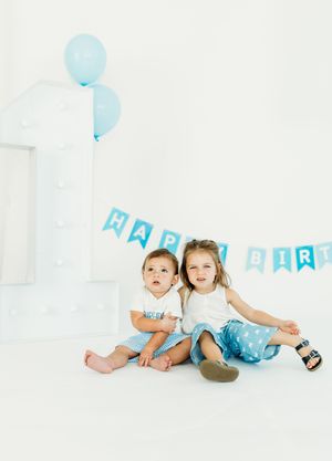 Three small children in light blue outfits play and pose together on a white backdrop with blue balloons and party decor.