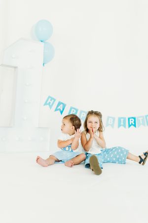Two children lying on a white floor with blue birthday decorations and balloons in a playful birthday photoshoot.