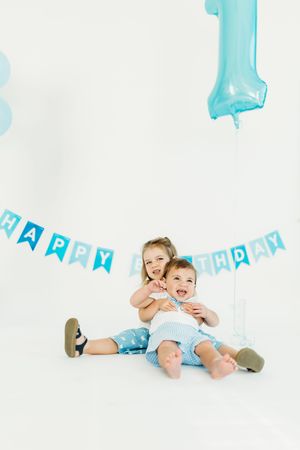 Two kids having fun during a birthday photo session with blue decorations and balloons in the background.