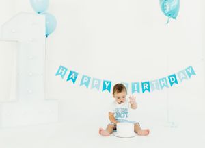 Baby boy in white shirt sits on floor with blue birthday decorations and balloons in background.