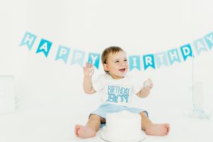 Baby in white onesie celebrates first birthday with blue banner decorations and cake smash session.