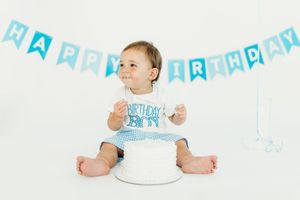 Baby wearing white shirt sits near birthday cake with blue banner decorations above.