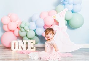 Baby girl in pink dress celebrates first birthday with pastel balloons and ONE sign backdrop.
