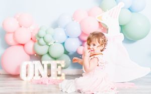 A baby girl in a pink dress celebrates her first birthday with colorful balloons and an ONE marquee sign.