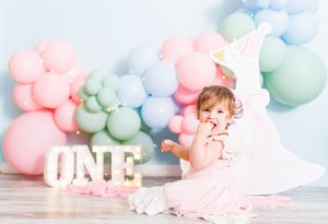 Baby girl in pink dress celebrates first birthday with colorful balloon garland and ONE marquee letters.