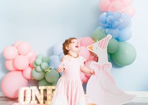 Toddler girl in pink dress laughing at first birthday party with colorful balloon garland and ONE sign.