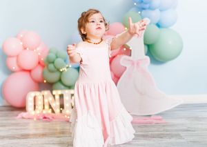 Toddler girl in pink dress reaches out at first birthday party with colorful balloon decorations and ONE sign.