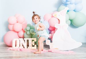 Toddler girl celebrates first birthday with colorful balloons, ONE marquee letters, and unicorn decor on pastel background.